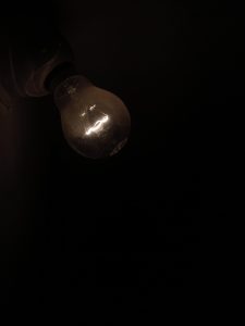 A single light bulb glows faintly in a dark space. Its warm filament softly breaks the surrounding darkness. The empty background adds a quiet, thoughtful mood.