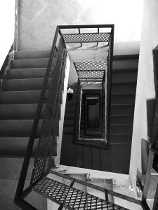 Black and white photograph of a multi-storey stairwell viewed from above, showing repeating geometric patterns of stairs and railings.