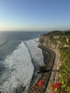 A sweeping coastal cliff curves along the shoreline as powerful waves meet the sea wall below, framed by lush greenery and a clear blue sky.