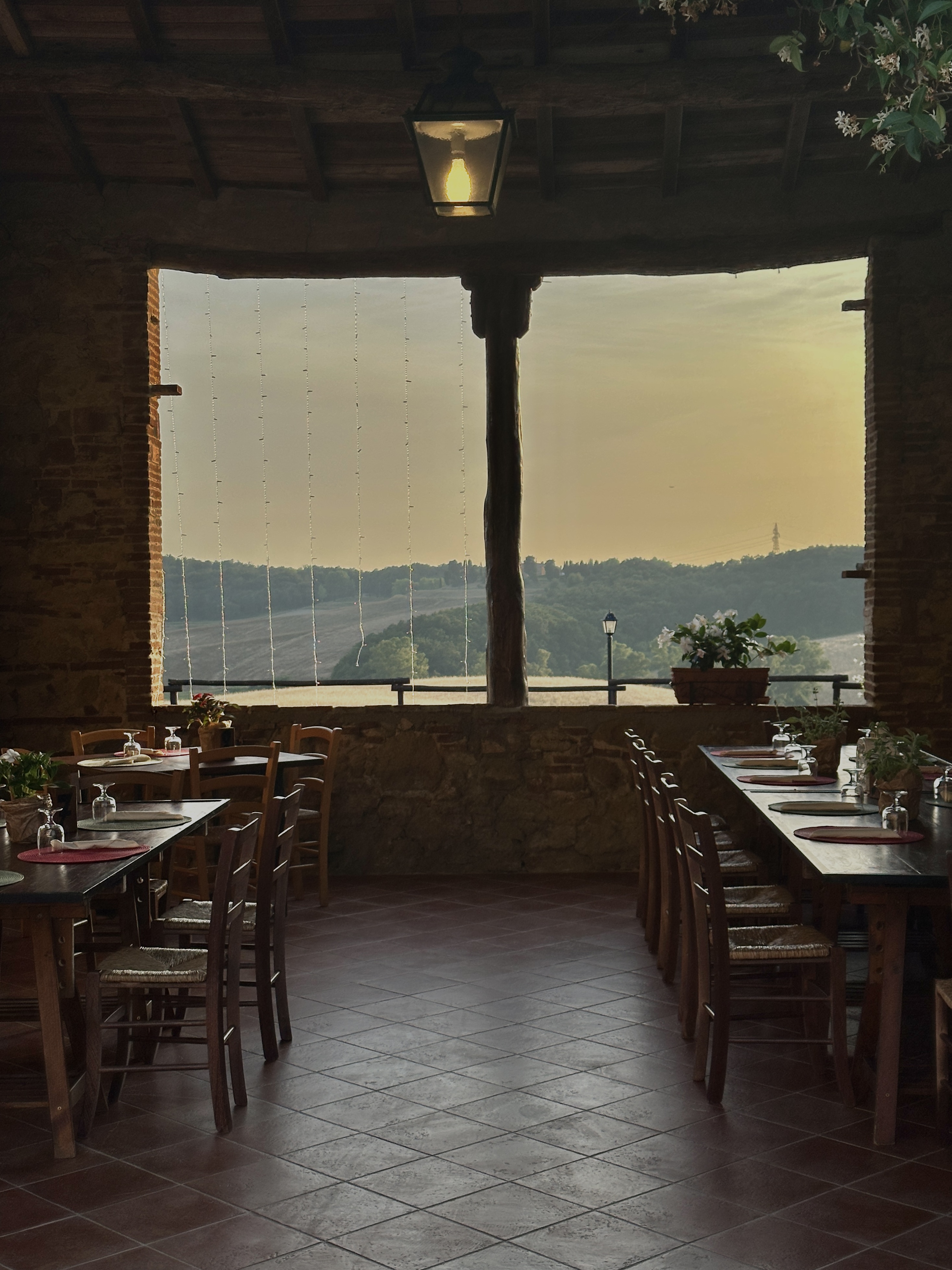 A cozy restaurant interior featuring wooden tables and chairs arranged neatly in front of a large window that offers a view of rolling hills.