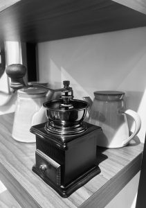 A black and white photo captures a vintage-style manual coffee grinder sitting next to ceramic pots on a wooden shelf.
