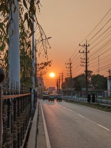 The image shows a busy street at sunset, with the sun glowing low on the horizon. There are vehicles, including small auto rickshaws and buses, driving down the road. Power lines on both sides of the road, and a few pedestrians walking. Trees and buildings are visible on either side of the road.