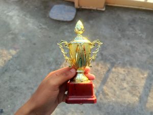 A person is holding a small golden trophy with decorative elements, including ornate leaf designs, and a red base.