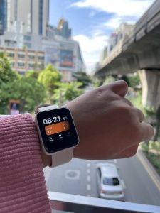A close-up of a smartwatch displaying the time as 08:21:03 along with health metrics, set against a blurred background of a city street with buildings and trees.