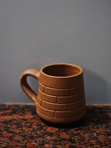 A handcrafted clay mug sits on a granite countertop in Perumanna, Kozhikode. The cup&rsquo;s brick-style design and warm, earthy tone stand out beautifully against the plain background, giving it a simple and rustic look. 