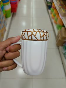 A person is holding a white coffee mug with a textured, golden decorative band around the top.