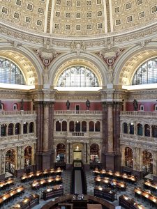 Grand historic library interior with ornate dome ceiling, marble columns, and reading desks arranged symmetrically.