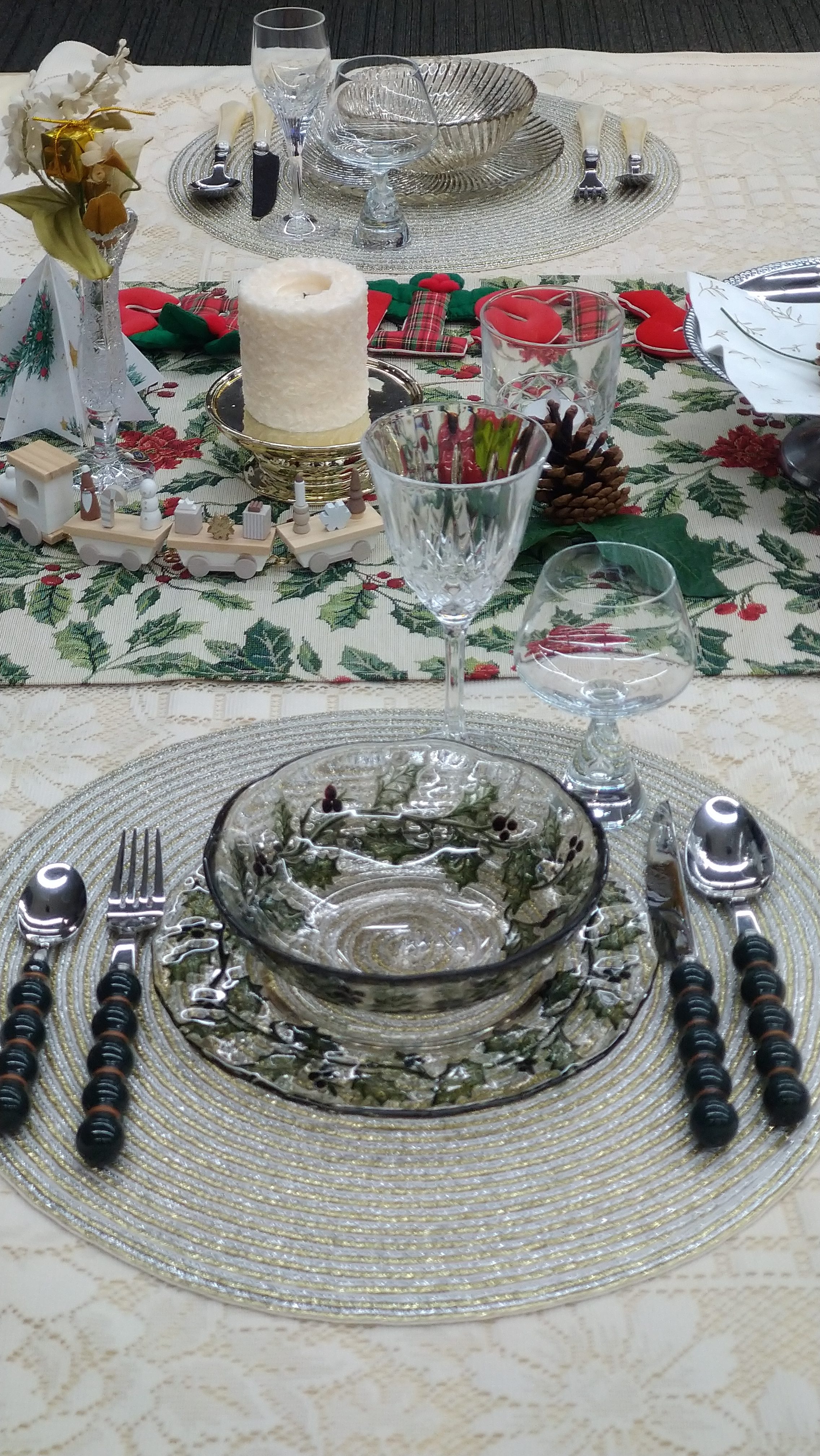 A nicely arranged dining table with a patterned tablecloth, a white candle, glasses, and dishes. The plates have green leaf designs, and there are small wooden items and pinecones on the table.