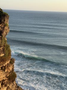Gentle ocean waves roll toward rugged coastal cliffs, bathed in soft golden light under a calm, open horizon.
