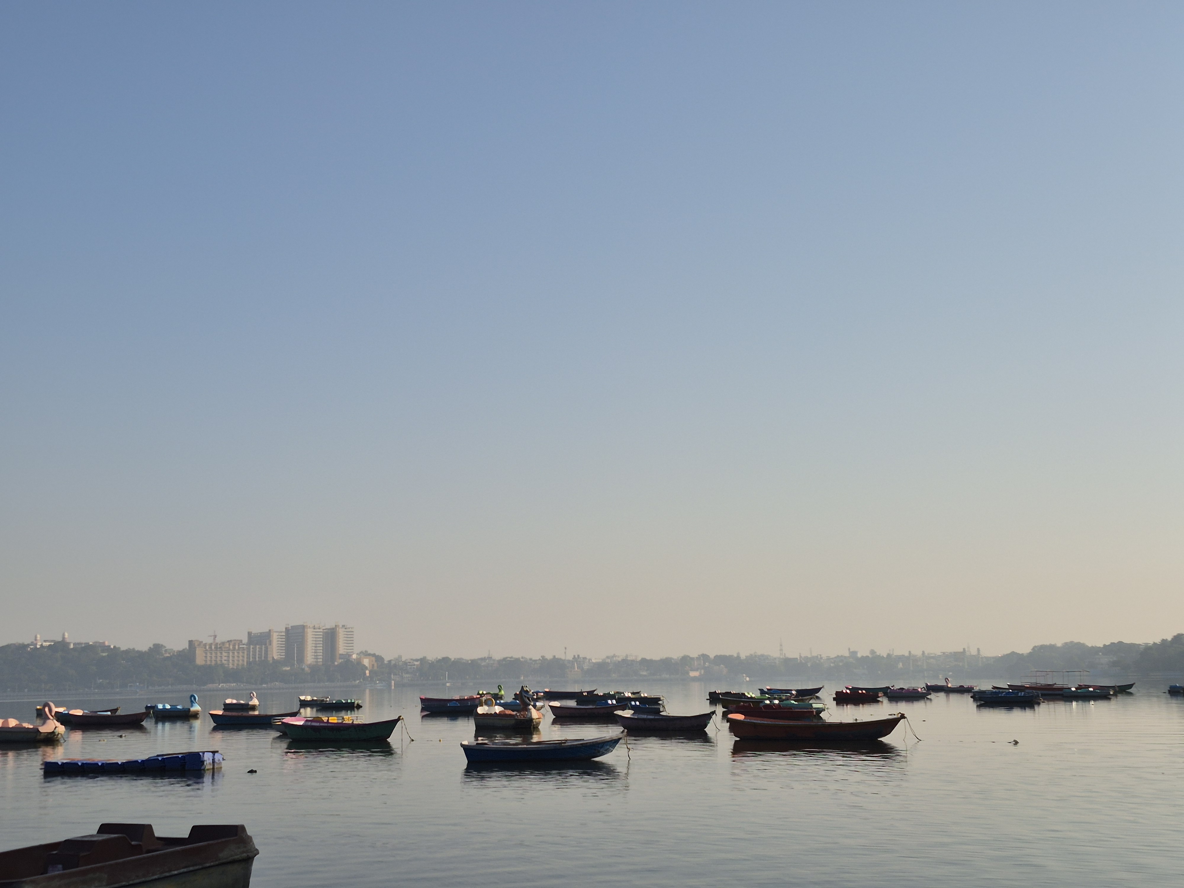 This image captures a serene, wide-angle view of a calm lake at what appears to be dawn or a hazy morning. The composition is defined by a large, clear sky that transitions from a pale blue to a soft, misty glow near the horizon.The overall mood is peaceful and rhythmic, highlighting the natural lake environment and the urban skyline beyond.
