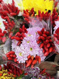 A close-up of colorful floral arrangements featuring bright red chili peppers mixed with pink and white chrysanthemums, alongside vibrant yellow and purple flowers. 
