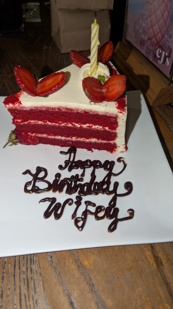 A slice of cake. The cake is bright red and the frosting is cream colored. On top are three strawberries cut so they’re open like clam shells.  Also a single candle stands in a bit of frosting. The cake is on a white board and on the board is written in icing “Happy Birthday Wifey”.