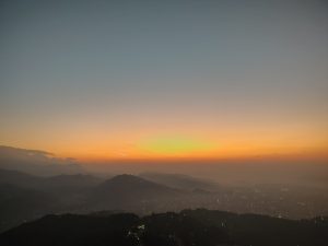A panoramic view of a sunset over a mountainous landscape. The sky is painted in gradient hues of orange, pink, and blue, transitioning from the warm colors of sunset to the cooler tones of twilight.