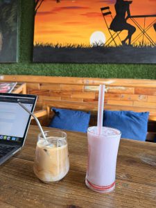 A cozy caf&eacute; setting featuring a wooden table with two drinks. On the left, a glass with a brown iced coffee topped with frothed milk and a metal straw.