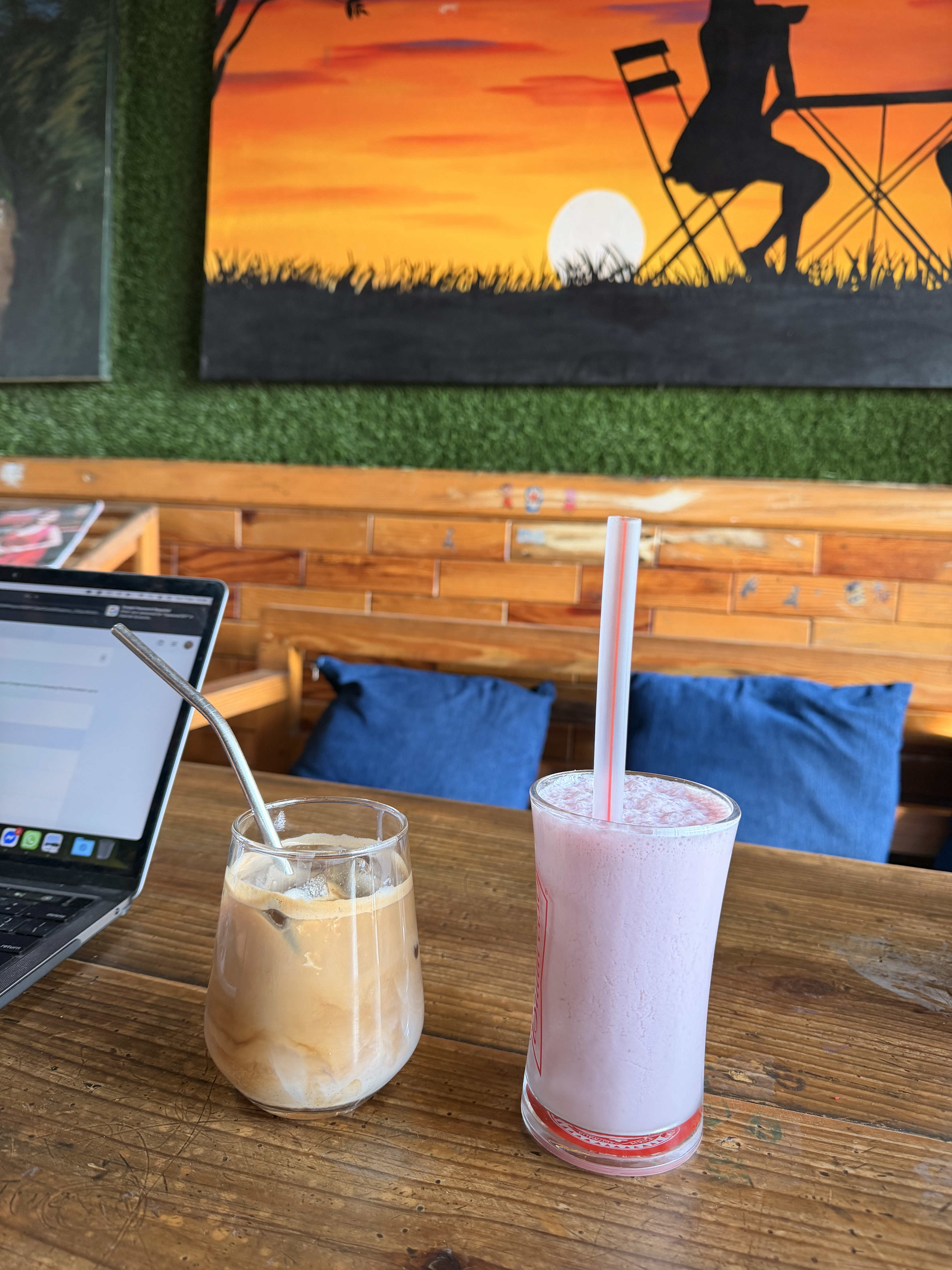 A cozy caf&eacute; setting featuring a wooden table with two drinks. On the left, a glass with a brown iced coffee topped with frothed milk and a metal straw.