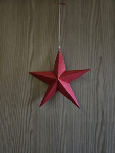 A handmade red paper star hanging against a simple wooden background. The clean design and soft light highlight the craft details, giving a calm, festive feel. This photo was taken in Kozhikode, Kerala, during the Christmas season. 
