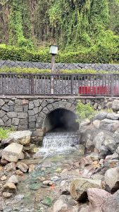 A serene scene featuring a stone bridge over a small, flowing stream. Water cascades through an arched opening beneath the bridge, creating a gentle waterfall effect.