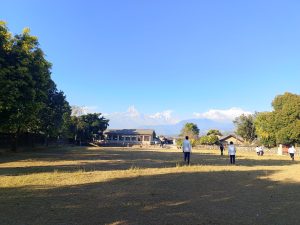 A picture of boys playing ball games in the grassy playground, which is in front of small one-story houses, some trees and a view of the mountain under the clear blue sky.