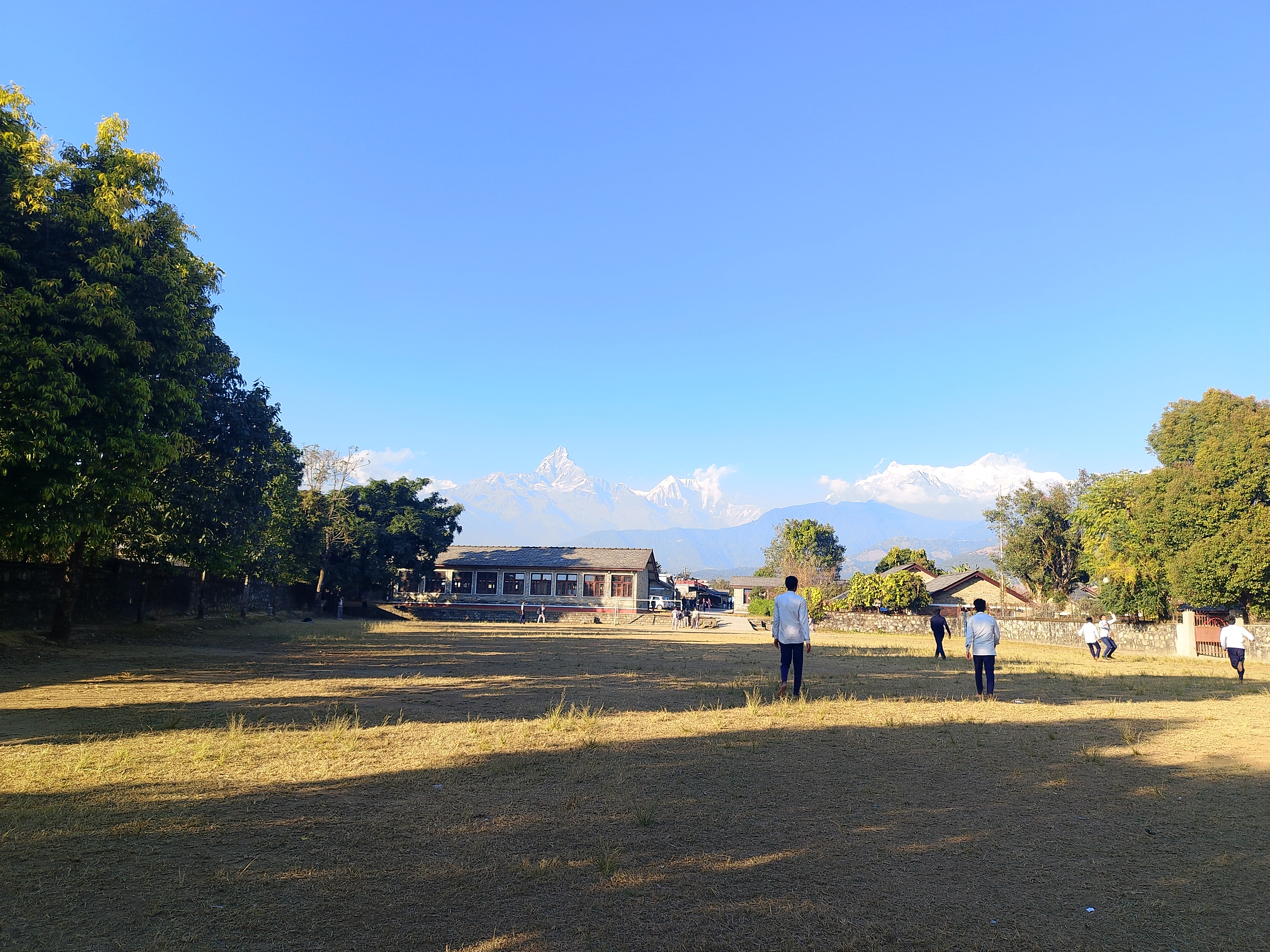 A picture of boys playing ball games in the grassy playground, which is in front of small one-story houses, some trees and a view of the mountain under the clear blue sky.