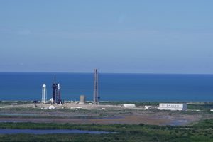 An aerial photo of SpaceX’s launch pad at NASA’s Kennedy Space Center, with the clear blue Atlantic Ocean in the background. The tall rocket support structure stands in the center.