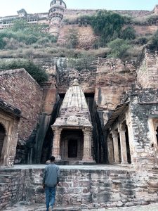 An ancient stone temple structure built into rocky surroundings. The walls show old carvings and weathered stone. A person is standing in front of the temple, adding scale and giving a historical, quiet feeling.