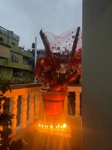 A red planter on a balcony railing holds decorative items wrapped in red netting and is illuminated by a row of small, bright oil lamps (diyas) in the foreground.
