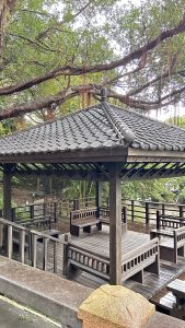 A wooden gazebo with a traditional tiled roof is surrounded by lush greenery.