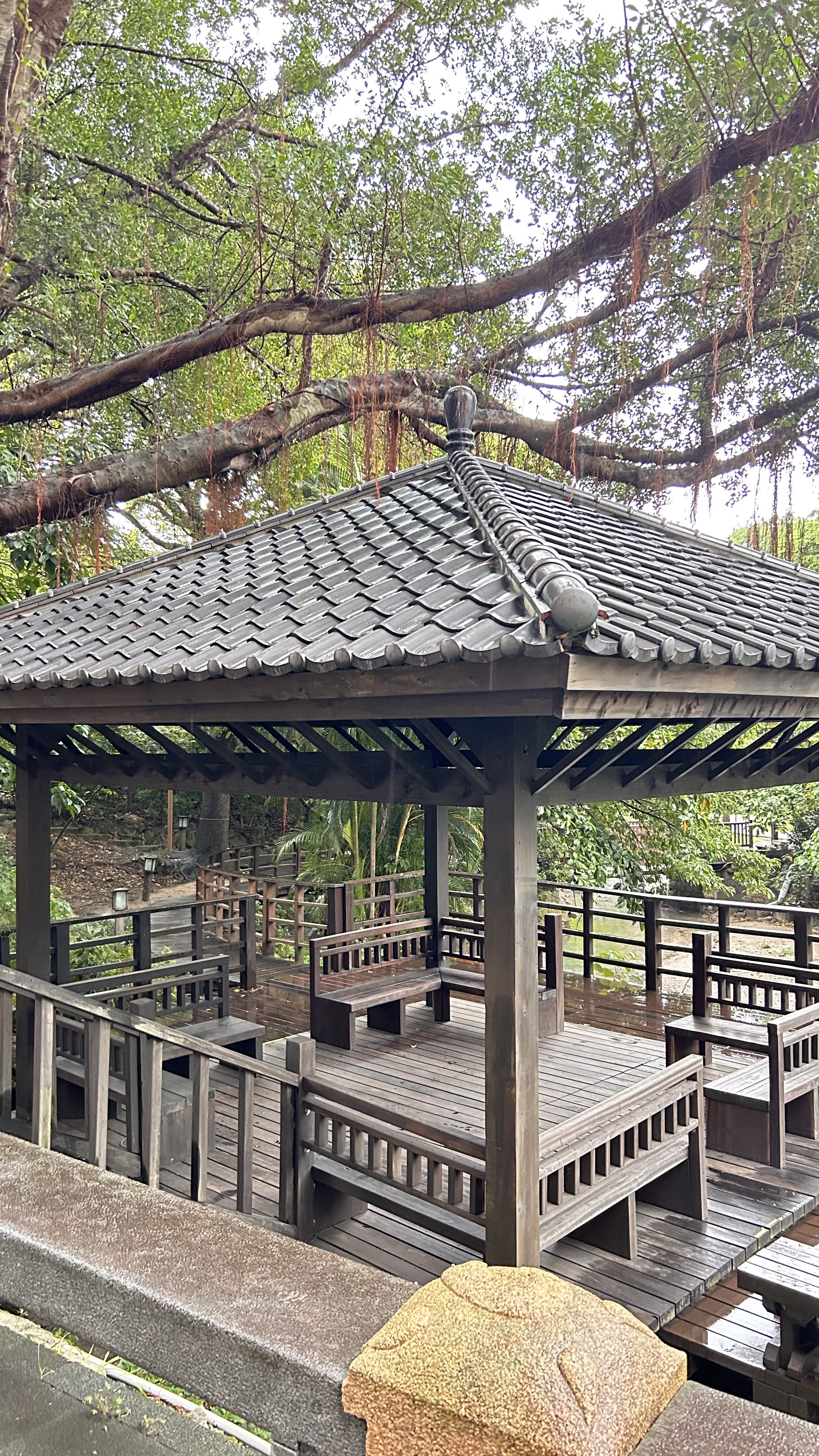 A wooden gazebo with a traditional tiled roof is surrounded by lush greenery.