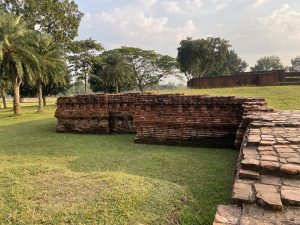 Ancient brick ruins on grassy terrain with palm and leafy trees in the background, under a partly cloudy sky, creating a serene, historic atmosphere.