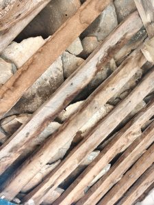 A close-up view of a rustic roof structure, featuring wooden beams laid across a foundation of irregularly shaped stones. 