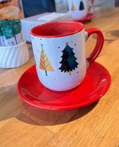 A festive cup adorned with illustrations of two Christmas trees, one in green and the other in gold, sits on a bright red saucer atop a wooden table.