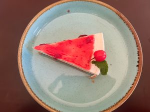 A slice of strawberry cheesecake with red fruit topping, a cherry, and a mint leaf sits on a light blue plate.