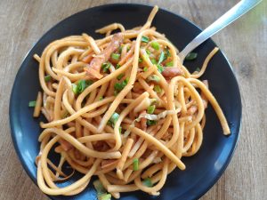 A black plate filled with stir-fried noodles, garnished with chopped green onions and small pieces of carrots.