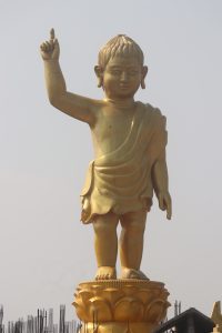Baby Buddha Statue of Lumbini Temple. The Buddha is cast as an infant, standing with the right arm raised and the pointer finger pointing to the sky. It’s made of a yellow metal and is many times larger than an adult human.
