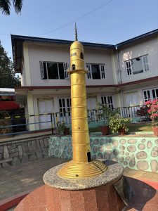 A tall, golden tower with a pointed spire stands prominently in front of a building.