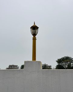 A tall, decorative wall lamp with a glass enclosure sits atop a yellow pole against a gray sky