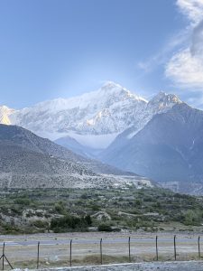 A panoramic view of snow-capped mountains under a blue sky, with the foreground showcasing a barren landscape and a barbed wire fence separating it from the mountains.