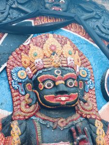 A colorful and detailed close-up of a statue depicting a deity, featuring a distinct face with large, vivid eyes and a strong expression. 