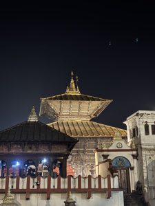 A night view of a historical temple complex, featuring a prominent pagoda-style roof with intricate wooden carvings and golden accents. 
