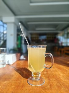 
A clear glass mug with a handle sits on a wooden table, containing a light yellow beverage.