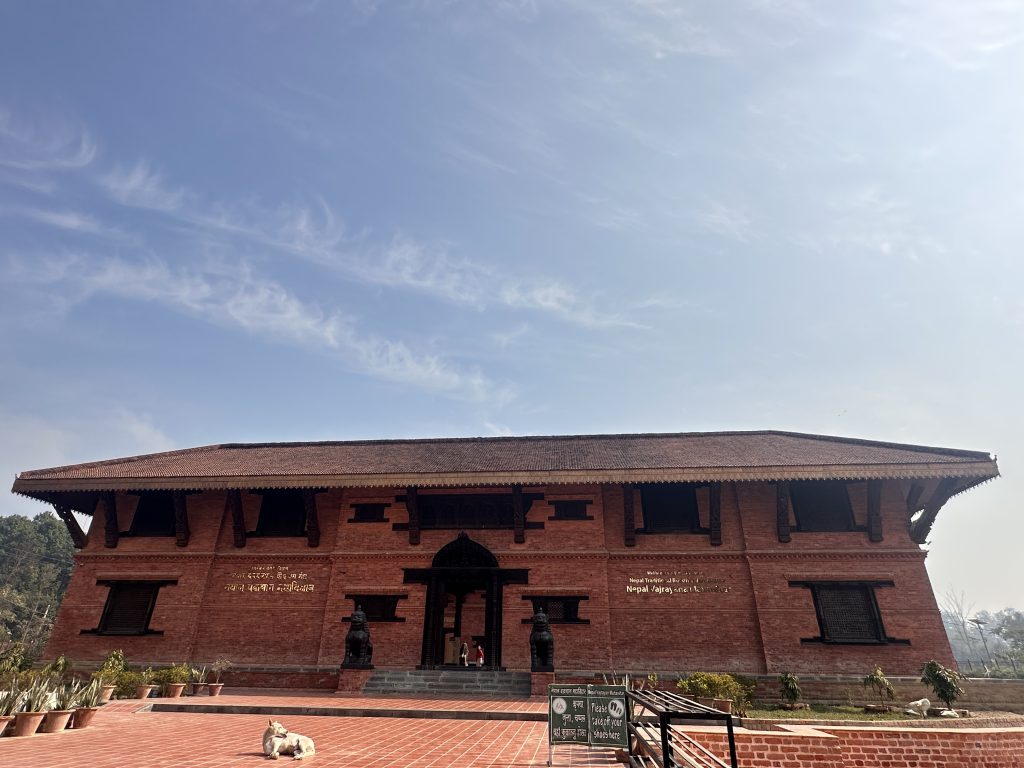 Monestry in Nepal. A large, traditional red brick building with a sloping roof is shown. The architecture features prominent wooden beams and windows, typical of Nepalese style. In front of the building, there are a couple of stone statues and a dog lying on the tiled walkway.
