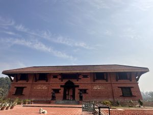 Monestry in Nepal. A large, traditional red brick building with a sloping roof is shown. The architecture features prominent wooden beams and windows, typical of Nepalese style. In front of the building, there are a couple of stone statues and a dog lying on the tiled walkway. 
