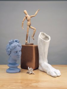 A photograph of a still life that includes the head of Aphrodite from Knidos in a pale, neutral, indigo-gray, a small wood mannequin posed in motion of a leap on top of a yoga cork block, a bone from an animal's neck, and a plaster cast foot on top of a wooden table and gray background.
