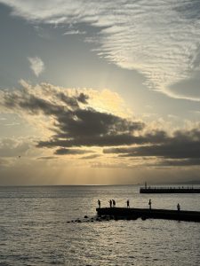 People standing on a pier by the sea at sunset under a cloudy sky.