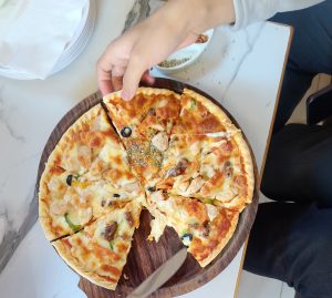 A wooden pizza board holds a freshly cut pizza topped with chunks of chicken, black olives, green peppers, and melted cheese.