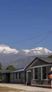 A scenic view featuring snow-capped mountains against a clear blue sky. In the foreground, a single-story stone building with a gray roof and red accents is visible.