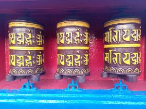 Three colorful prayer wheels with yellow script are mounted on blue supports against a red background, suggesting a place of worship or meditation.