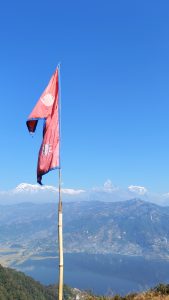 
A flag of Nepal flutters at the top of a mountain against a clear blue sky. In the background, majestic snow-capped peaks are visible, while a tranquil lake reflects the surrounding landscape below. The scene showcases a blend of natural beauty and cultural significance.