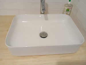 A bathroom style sink, very modern, with a flat bottom sitting on top of a wooden table.  It the top right is a bottle of hand soap.