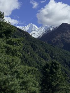 A scenic view of snow-capped mountains rising above lush green hills, with a clear blue sky and fluffy white clouds in the background. 